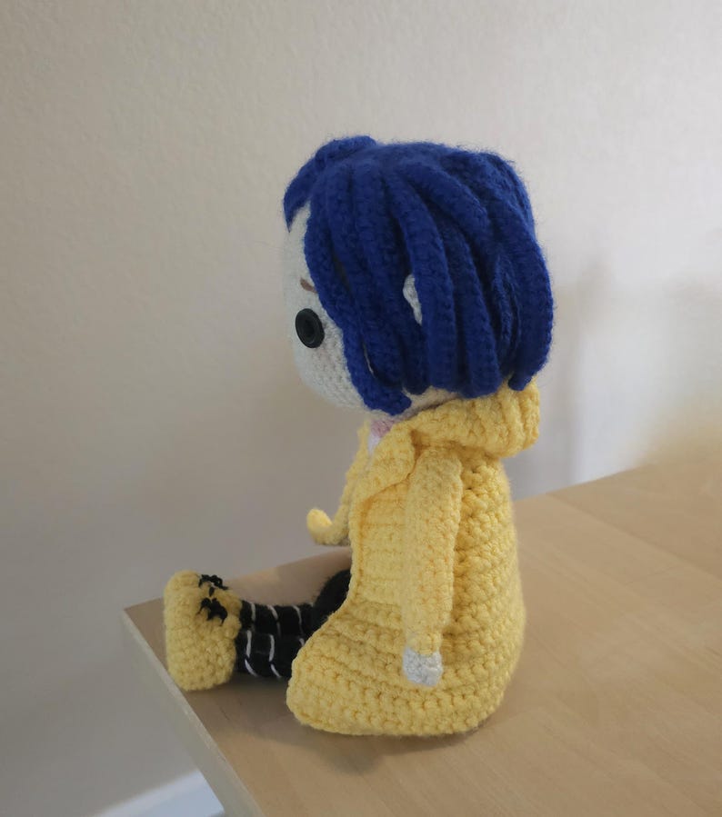 May include: Crocheted doll featuring a character with blue hair, a white face, and a yellow hooded jacket. The doll is seated, with black and white striped pants and black shoes. The doll is handmade and appears to be a character from a movie.