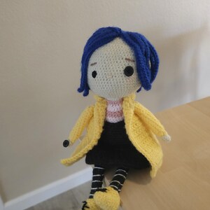 May include: Crocheted doll of a character with blue hair, wearing a yellow coat, black skirt, and striped socks. The doll has black button eyes and yellow shoes. The doll is sitting on a wooden surface.