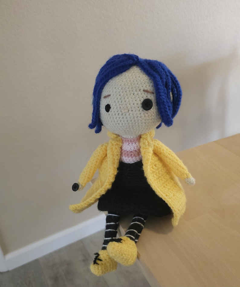 May include: Crocheted doll of a character with blue hair, wearing a yellow coat, black skirt, and striped socks. The doll has black button eyes and yellow shoes. The doll is sitting on a wooden surface.