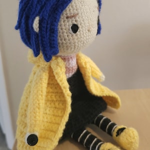May include: Crocheted doll with blue hair, wearing a yellow coat, black skirt, and striped leggings. The doll has a button on the coat and yellow shoes. The doll is sitting on a wooden surface.