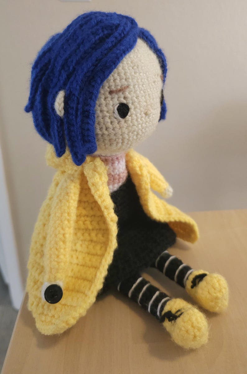 May include: Crocheted doll with blue hair, wearing a yellow coat, black skirt, and striped leggings. The doll has a button on the coat and yellow shoes. The doll is sitting on a wooden surface.
