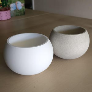 May include: Two spherical planters. One is smooth white, the other is speckled gray. Both have a rounded shape with a wide opening. These decorative planters are suitable for small plants or succulents.