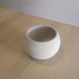 May include: A speckled, light grey, spherical bowl. The bowl has a smooth, rounded exterior and a wide opening. The interior is a lighter shade of grey. The bowl is on a light brown wooden surface.