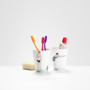 Peut inclure: Deux porte-brosses à dents en céramique blanche avec des illustrations de personnes en maillot de bain. Un porte-brosse a un homme avec un canard, l'autre a une femme les bras tendus. Trois brosses à dents sont dans le porte-brosse de gauche, une orange, une rose et une violette. Un tube de dentifrice est dans le porte-brosse de droite.