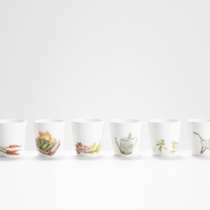 May include: Six white ceramic mugs with different garden-themed illustrations. The mugs feature images of carrots, a watering can, a box of vegetables, a bunch of flowers, a sprig of herbs, and a white rabbit.