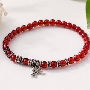 May include: A beaded bracelet featuring small, translucent deep red circular beads interspersed with ornate, textured silver-toned metal spacers. A small, detailed silver-toned metal dragonfly charm hangs from the center of the bracelet.