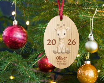 Può includere: Un addobbo natalizio ovale in legno con un'illustrazione stampata di una giraffa neonata seduta. I numeri 20 e 26 sono stampati su entrambi i lati della giraffa. Sotto la giraffa, il nome Oliver e il testo Baby's First Christmas.