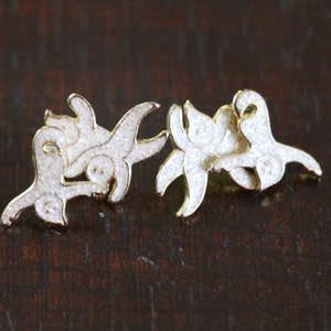 May include: A pair of gold-tone earrings with white glitter accents. The earrings are shaped like a stylized floral design.