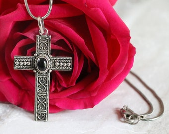 Op de afbeelding: Een zilveren ketting met een gedetailleerde kruis hanger met een zwarte edelsteen. De ketting ligt op een rode roos.