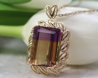 Può includere: Collana con pendente in oro con una gemma rettangolare bicolore nei toni del viola e del giallo. Il pendente è incorniciato da un bordo dorato decorativo e pende da una catena sottile. La collana è presentata su una superficie bianca.