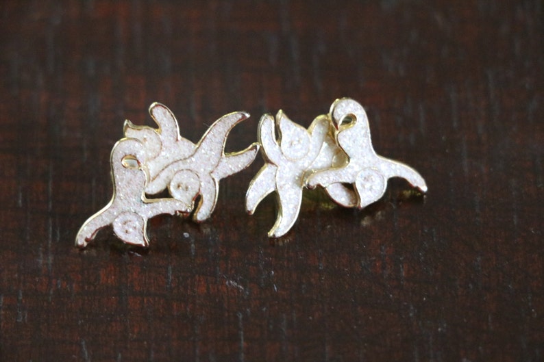 May include: A pair of gold-tone earrings with white glitter accents. The earrings are shaped like a stylized floral design.
