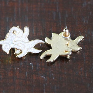May include: A pair of gold-tone earrings with a white enamel design. The earrings are shaped like a stylized bird with its wings spread.