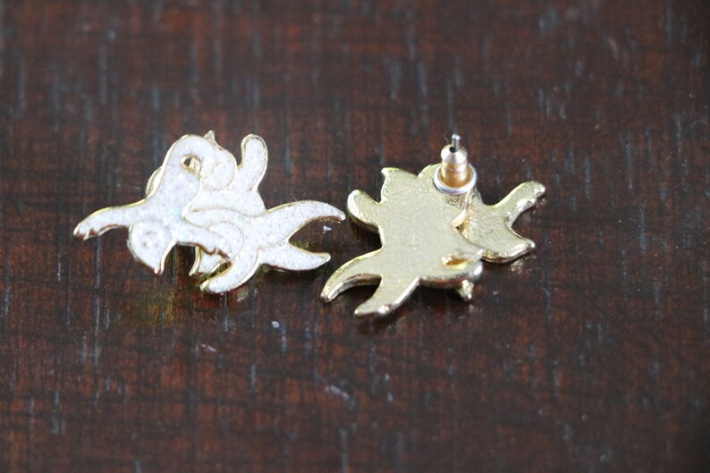 May include: A pair of gold-tone earrings with a white enamel design. The earrings are shaped like a stylized bird with its wings spread.