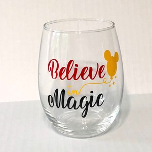 May include: A clear glass wine glass with the text "Believe in Magic" in red and black lettering. A yellow balloon with a Mickey Mouse head is above the text.