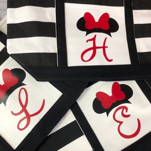 K&ouml;nnte beinhalten: Drei schwarze und wei&szlig;e gestreifte Einkaufstaschen mit einem roten Schleifen- und schwarzen Ohren-Design. Die Taschen haben eine schwarze Tasche auf der Vorderseite mit einem roten Buchstabenmonogramm. Die Buchstaben sind "H", "J" und "E".