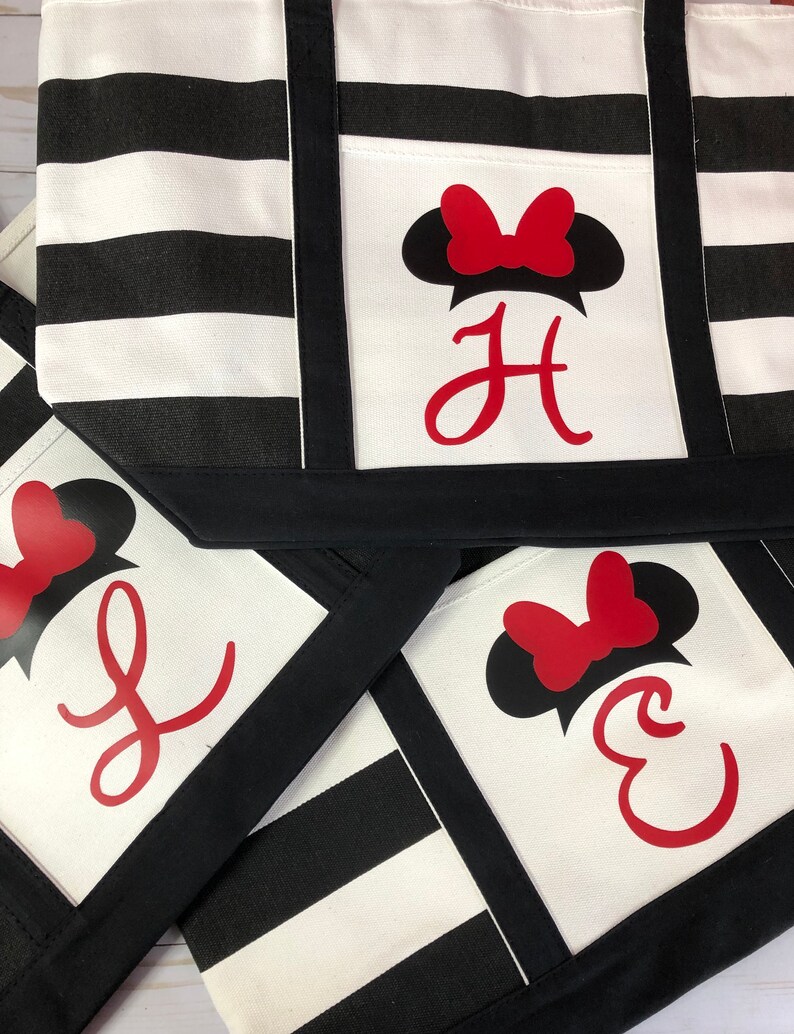 K&ouml;nnte beinhalten: Drei schwarze und wei&szlig;e gestreifte Einkaufstaschen mit einem roten Schleifen- und schwarzen Ohren-Design. Die Taschen haben eine schwarze Tasche auf der Vorderseite mit einem roten Buchstabenmonogramm. Die Buchstaben sind "H", "J" und "E".