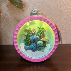 May include: A pink and green Christmas ornament featuring a scene with Stitch and another character. The ornament has a silver top and a string for hanging. The interior scene includes two small Christmas trees and string lights.