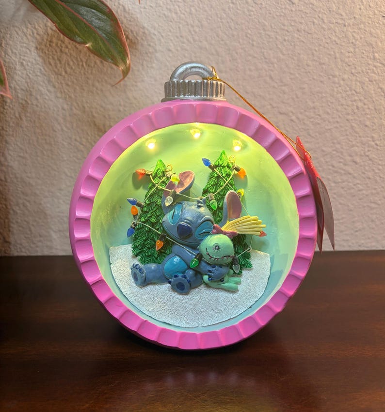 May include: A pink and green Christmas ornament featuring a scene with Stitch and another character. The ornament has a silver top and a string for hanging. The interior scene includes two small Christmas trees and string lights.