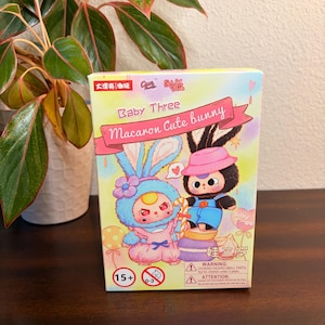 May include: A colorful box featuring the text "Baby Three Macaron Cute bunny" with illustrations of two cartoon bunnies. One bunny is blue, and the other is black. The box has a warning label and is set against a neutral background.