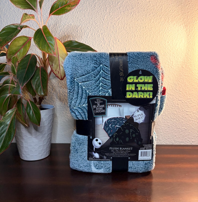 May include: A plush blanket featuring a design from The Nightmare Before Christmas. The blanket is blue with a tree and web pattern, and the packaging states it glows in the dark and is super soft. The blanket is wrapped with a black band.