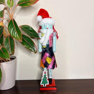 May include: A wooden nutcracker doll in the style of Sally from The Nightmare Before Christmas, wearing a Santa hat and a white scarf. The doll has a light blue face and arms, a patchwork skirt, and stands on a red base with a Christmas tree.