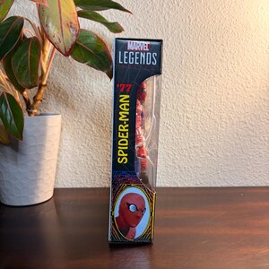 May include: Marvel Legends '77 Spider-Man action figure in its original packaging. The box is black with yellow and red accents, featuring the Marvel Legends logo and the Spider-Man name. The figure is visible through a clear plastic window.
