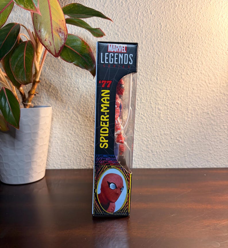 May include: Marvel Legends '77 Spider-Man action figure in its original packaging. The box is black with yellow and red accents, featuring the Marvel Legends logo and the Spider-Man name. The figure is visible through a clear plastic window.