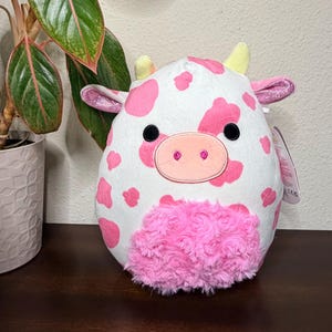 May include: A plush cow toy with a white body and pink spots. The cow has a pink snout, black eyes, and pink ears with glitter accents. The bottom of the cow is covered in pink, fluffy material. A tag is attached.