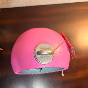 May include: A bright pink, dome-shaped ornament with a silver metal cap. A small red tag is attached with gold string. The bottom edge is a light blue color. The ornament is likely for holiday or seasonal decor.