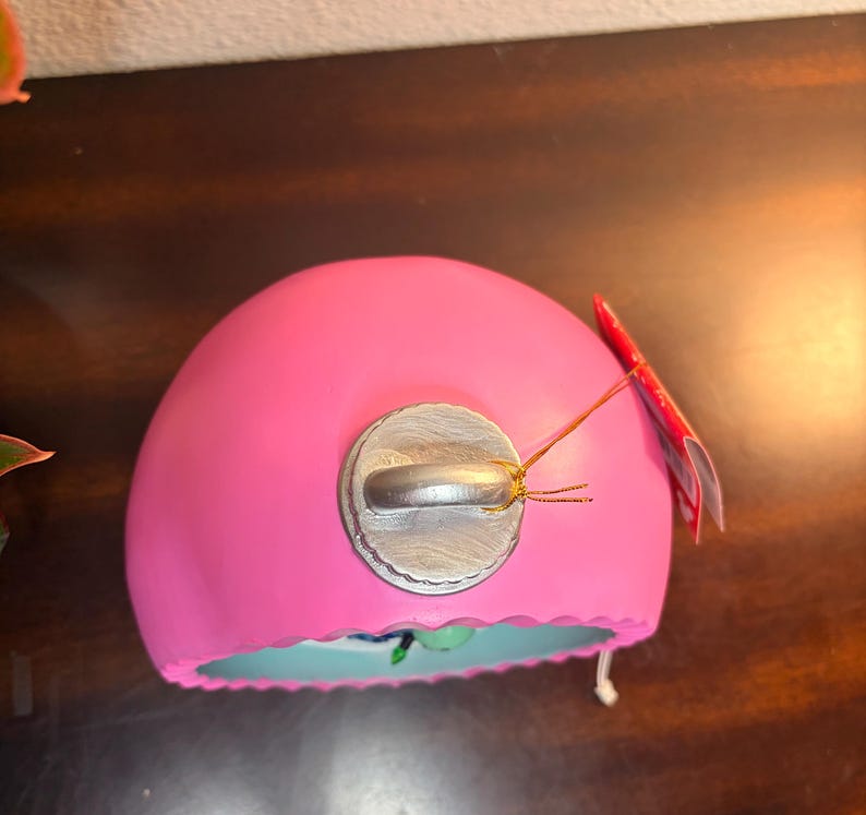 May include: A bright pink, dome-shaped ornament with a silver metal cap. A small red tag is attached with gold string. The bottom edge is a light blue color. The ornament is likely for holiday or seasonal decor.