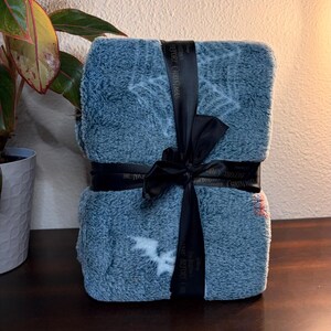 May include: A folded, blue blanket with white snowflake and bat patterns, tied with a black ribbon. The ribbon has text that reads "The Nightmare Before Christmas". The blanket is on a dark wooden surface.
