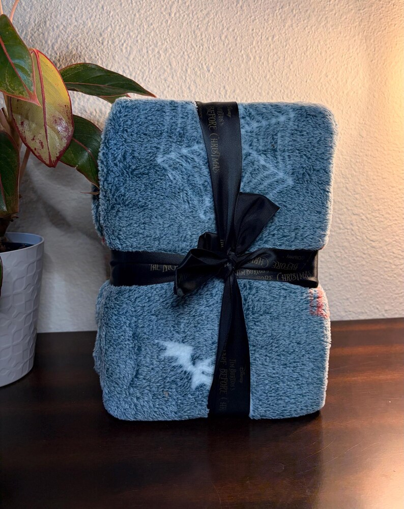 May include: A folded, blue blanket with white snowflake and bat patterns, tied with a black ribbon. The ribbon has text that reads "The Nightmare Before Christmas". The blanket is on a dark wooden surface.