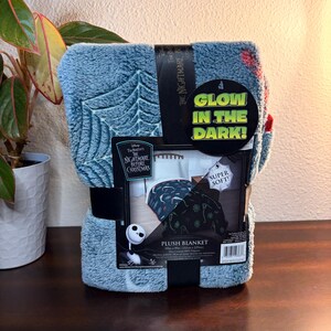 May include: A blue plush blanket featuring a web design and the text "The Nightmare Before Christmas." The blanket is packaged with a black band and a tag that reads "Glow in the Dark!" and "Super Soft!" The blanket measures 60 x 90 inches.
