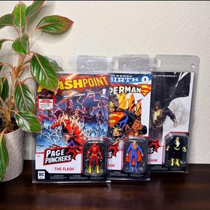 May include: Comic books and action figures featuring DC Comics characters. The Flash comic book is in the foreground, with a Flash action figure below. Superman and Black Adam figures are also visible, along with other comic books.