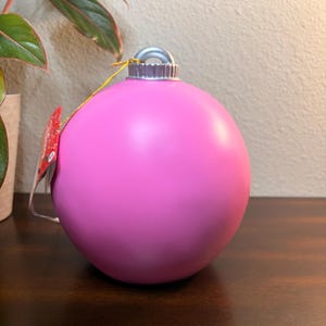 May include: A large, bright pink Christmas ornament with a silver cap and a gold string. The ornament is smooth and spherical, with a tag attached. The background includes a plant and a wooden surface.