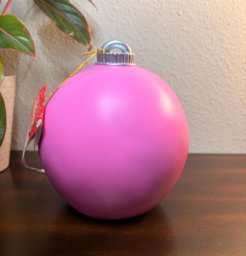 May include: A large, bright pink Christmas ornament with a silver cap and a gold string. The ornament is smooth and spherical, with a tag attached. The background includes a plant and a wooden surface.