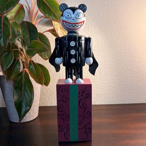 May include: A black and white wooden figurine of a vampire-like character with a wide, toothy grin, standing on a purple block with green and black swirl designs. The figurine has a black suit with white buttons and small black capes.