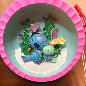 May include: A Christmas ornament featuring Stitch and a green alien character hugging. The ornament has a pink scalloped edge and a light blue interior with two Christmas trees and string lights. The characters are blue and green.