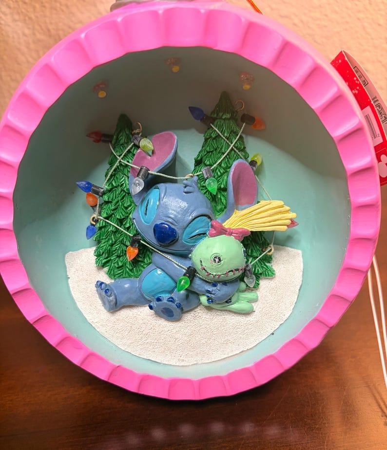 May include: A Christmas ornament featuring Stitch and a green alien character hugging. The ornament has a pink scalloped edge and a light blue interior with two Christmas trees and string lights. The characters are blue and green.