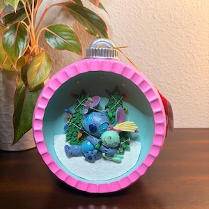 May include: A pink and blue Christmas ornament featuring Stitch and a green alien character. The ornament has a pink rim and a light blue interior with miniature Christmas trees and string lights. The characters are lying on a snowy surface.
