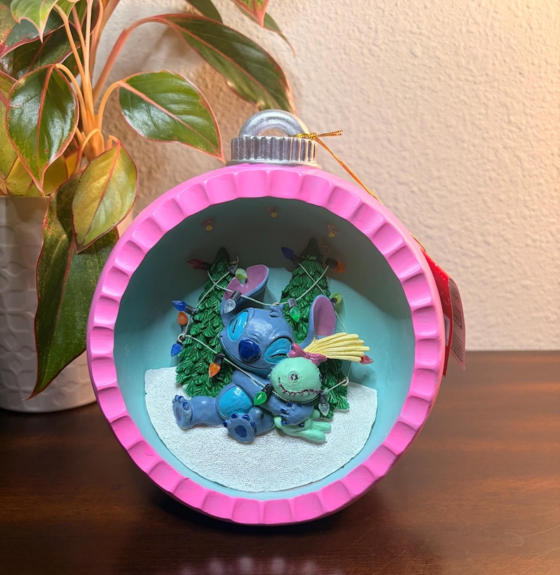 May include: A pink and blue Christmas ornament featuring Stitch and a green alien character. The ornament has a pink rim and a light blue interior with miniature Christmas trees and string lights. The characters are lying on a snowy surface.