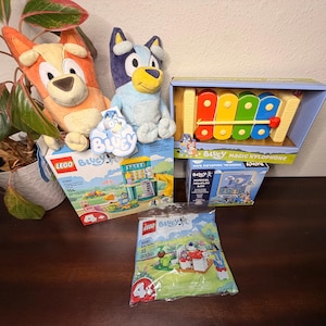 May include: A collection of Bluey-themed toys and games. Includes plush toys of Bluey and Bingo, a Bluey Magic Xylophone, and two Lego Bluey sets. The Lego sets are labeled with the number of pieces and age range.