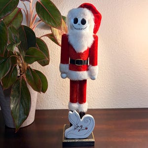 May include: A decorative wooden nutcracker in the style of Jack Skellington dressed as Santa Claus. The figure is red and white with a black belt and a white beard. A small wooden Zero figure is at the base.
