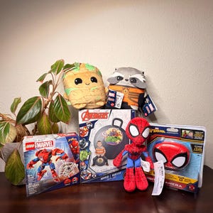 May include: A collection of Marvel-themed toys and merchandise, including a LEGO Marvel Spider-Man set, plush characters of Groot and Rocket, a Spider-Man plush doll, a Spider-Man digital camera, and an Avengers hopper ball.