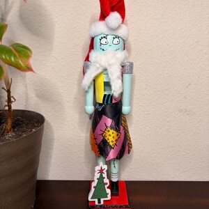 May include: A decorative nutcracker figure featuring a pale blue face with stitched details, wearing a red and white Santa hat and a white faux fur scarf. The body is dressed in a patchwork-style outfit with a small Christmas tree attached to the base.
