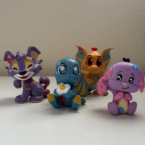 Puede incluir: Cuatro figuras coloridas de criaturas fantásticas. Las figuras incluyen una criatura morada con forma de gato, una criatura azul con forma de tortuga que sostiene una flor, una criatura naranja con forma de dragón y una criatura morada con forma de conejo.