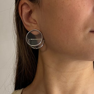 May include: Silver-toned, geometric earrings with a circular design. The earrings feature a layered, open-circle construction with a horizontal bar across the top. The jewelry is worn on the ear. The model is wearing a cream-colored tank top.
