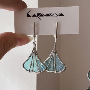 May include: Silver-tone earrings with a fan-shaped design. The earrings have a light blue and teal stained glass pattern, with a silver border. The earrings are displayed on a white card with the text "KARAVAVA GLASS ART".