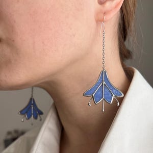 Puede incluir: Un par de pendientes de plata con colgantes de flores de vidrio azul. Las flores tienen un diseño de cinco pétalos y están suspendidas de una cadena de plata.