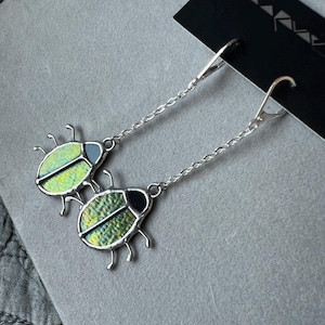 Puede incluir: Pendientes colgantes plateados con diseño de mariquita. Cada pendiente tiene un pequeño y detallado dije de mariquita con cuerpo verde y amarillo, detalles negros y contorno plateado. Los pendientes cuelgan de una cadena y un cierre de palanca.